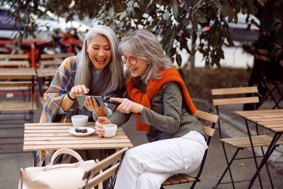 Positive senior lady with friend surf internet on smartphone in street cafe
